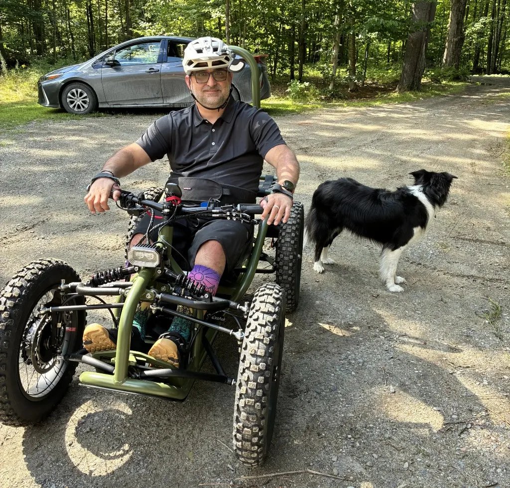 Yann in an Exoquad on a forest trail, with a dog standing beside the machine